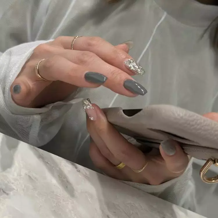 A imagem mostra unhas longas e ovais, pintadas em tons de cinza escuro e nude. Algumas unhas têm detalhes em glitter prateado, adicionando um toque de brilho. A pessoa usa anéis dourados finos nos dedos e segura uma bolsa bege. O fundo é uma superfície de mármore, complementando o visual sofisticado e moderno.


