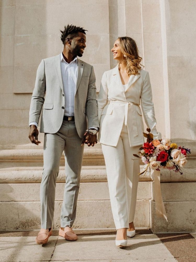 Mulher de pele clara usando conjunto de blazer e calça branca, segurando buquê e de mãos dadas com noivo de pele negra usando terno cinza