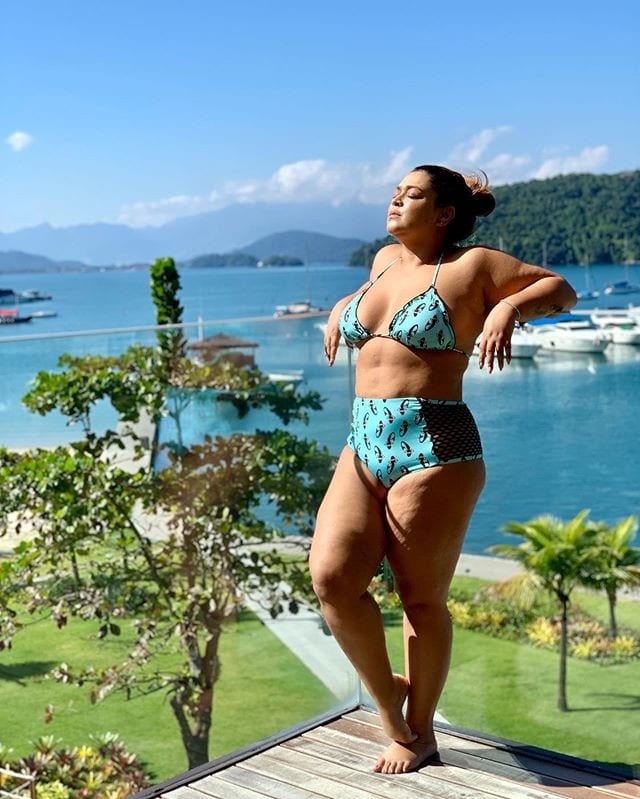 Cantora Preta Gil tomando sol de biquíni azul, apoiada em sacada de vidro com o mar repleto de barcos ao fundo.