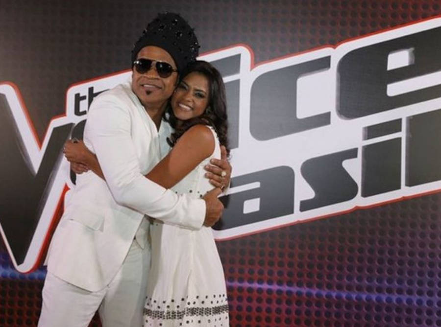 Foto de Lucy Alves e Carlinhos Brown na frente de uma placa do The Voice Brasil, ambos estão de roupa branca