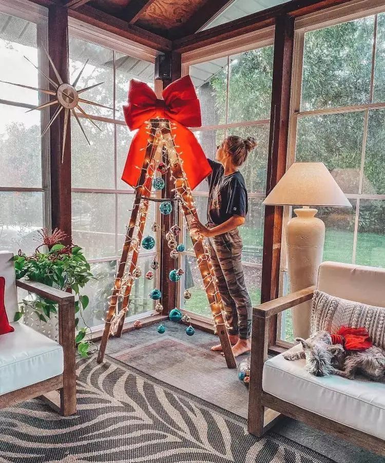 Uma mulher decora uma árvore de Natal feita com escada de madeira, iluminada por luzes e enfeitada com bolas coloridas. No topo, um laço vermelho. Ao lado, um cachorro descansa em uma poltrona. O ambiente é claro, com grandes janelas e plantas.


