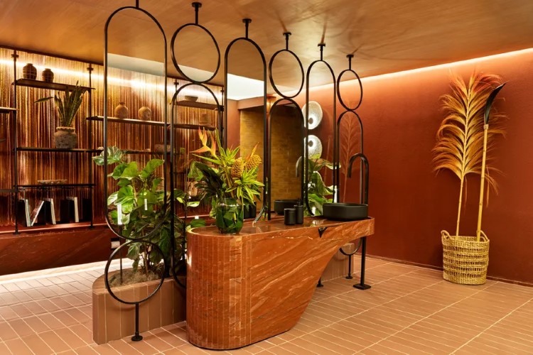 A imagem mostra um banheiro moderno com bancada de mármore marrom, pia preta, espelho oval, luminárias circulares metálicas, plantas tropicais e azulejos rosados no chão.

