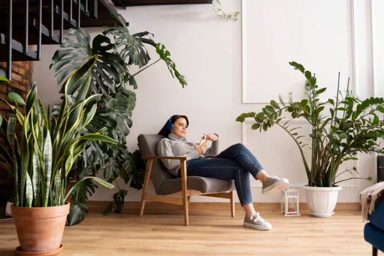Mulher de pele clara e cabelo escuro com fone de ouvido e segurando um celular. Ela está sentada em uma poltrona, disposta em um ambiente relaxante com plantas e em cores neutras, como branco e tom de madeira, abaixo de uma escada preta. A decoração contribui para a saúde mental