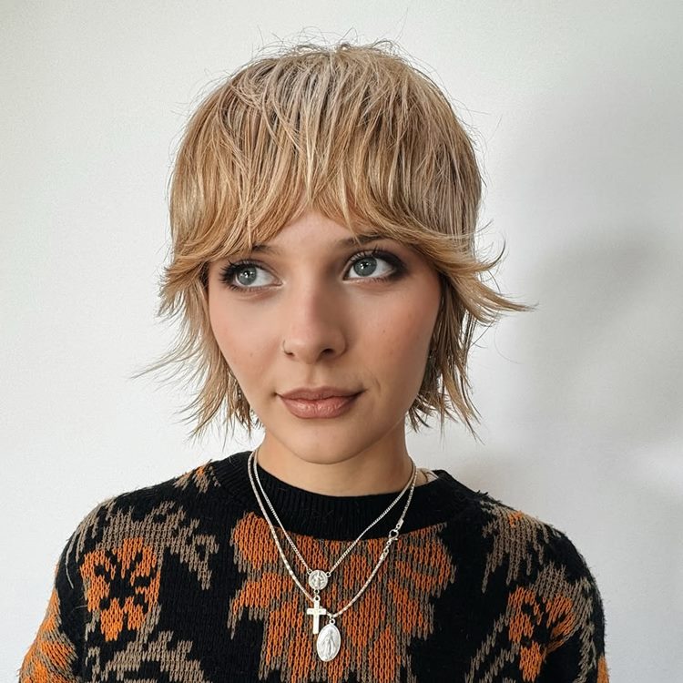 Jovem mulher com cabelo loiro curto e desfiado, usando suéter com estampa floral marrom e preta. Ela tem olhos expressivos, maquiagem suave e colares prateados. A expressão é serena, capturada contra um fundo branco, destacando um estilo moderno e despojado.


