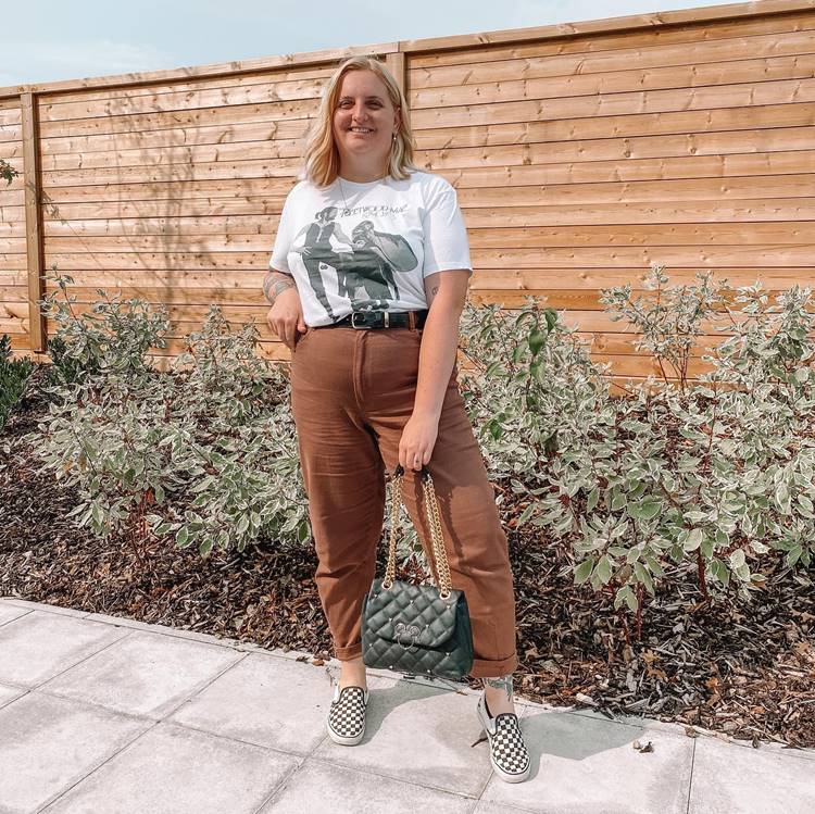 Garota loira com corpo mid size usando camiseta branca estampada, calça marrom com cinto preto e tênis quadriculado, em local com muro de madeira e plantas.