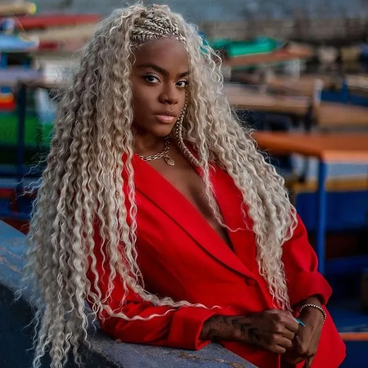 Mulher negra com roupa vermelha e trança nagô até metade da cabeça. Seu cabelo é longo e platinado. Ela está encostada em muro. O fundo está desfocado.