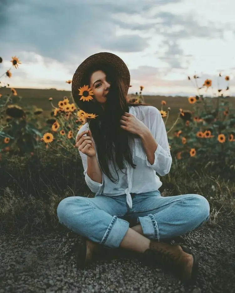 Frase para foto pensativa mulher com calça jeans, camisa branca chapéu de palha e bota de camurça segura girassol na mão Frase para foto pensativa