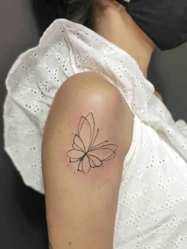 Tatuagem de borboleta com poucos traços, sendo eles finos e poucos detalhes. Feita no braço, próxima ao ombro, de uma mulher de pele clara vestindo uma blusa branca.