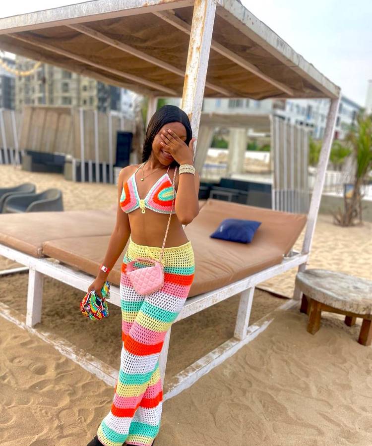 Uma mulher sorri timidamente, cobrindo o rosto, em uma barraca de praia. Ela usa um conjunto de crochê colorido, com um top estilo halter neck e calças de perna larga listradas em tons pastel.