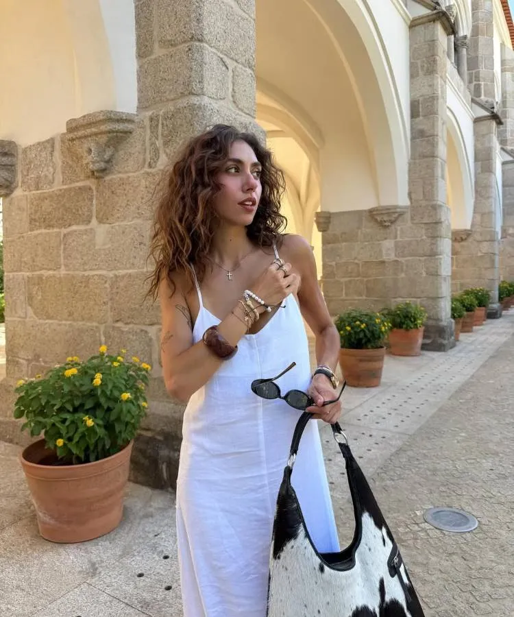 Mulher de cabelos cacheados e castanhos, usando um vestido branco, segurando óculos de sol e uma bolsa grande com estampa de vaca. Ela está em um pátio com arcos de pedra, olhando para o lado.