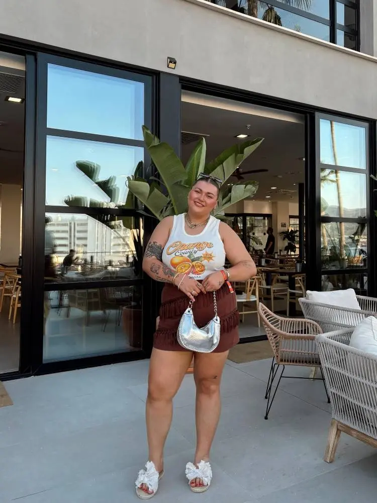 Uma mulher sorridente com cabelos escuros presos e tatuagens posa em frente a um restaurante. Ela veste um top branco com estampa de laranja, uma saia marrom de franjas, uma bolsa prateada e chinelos de palha com laços brancos.