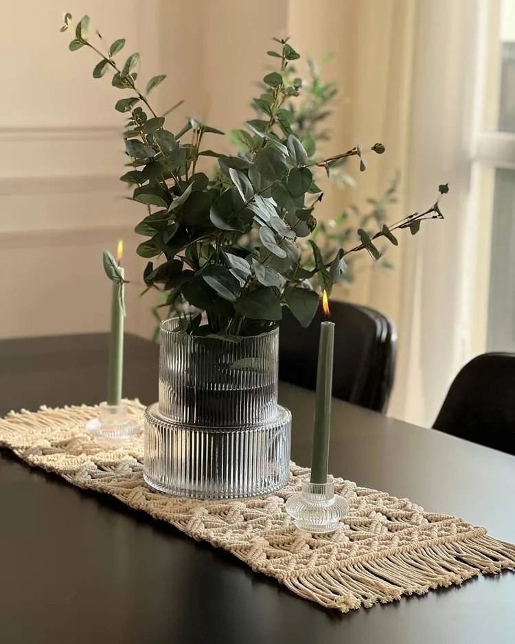 mesa com uma toalha de macramê bege no centro. Sobre a toalha, há um vaso de vidro transparente com folhagens verdes e duas velas verdes acesas em castiçais de vidro, uma de cada lado do vaso.