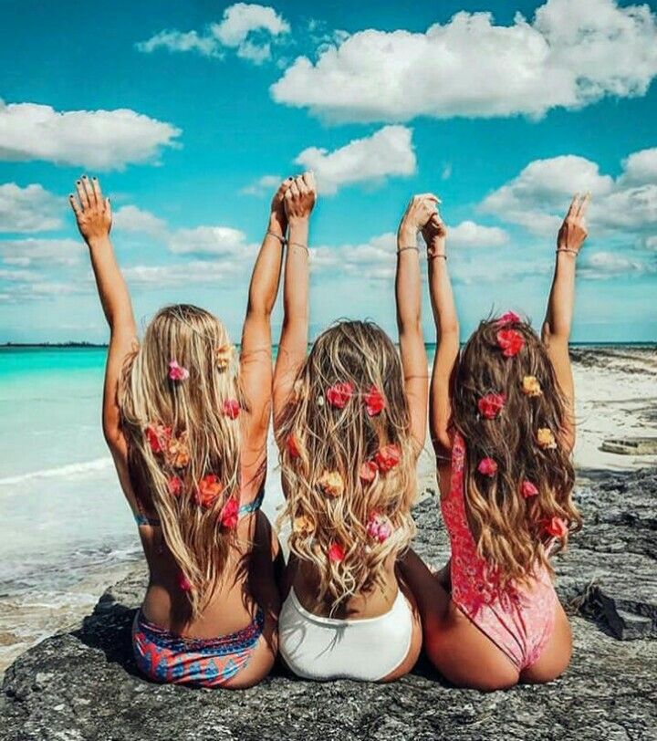 Três mulheres loiras de pele clara sentadas de costas em pedra à beira do mar com flores no cabelo 