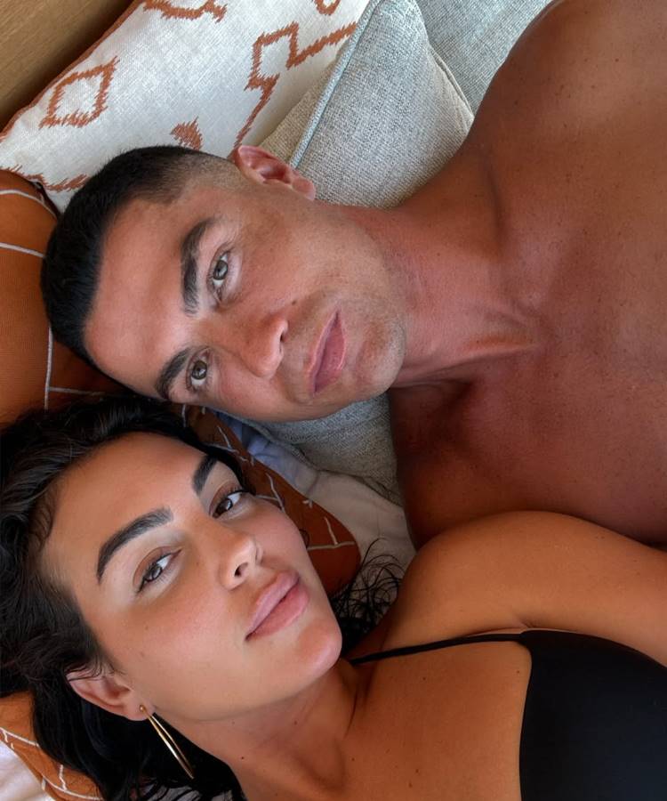 Cristiano Ronaldo e Georgina Rodríguez deitam lado a lado, com a cabeça no travesseiro. Os dois olham diretamente para a câmera, com expressões relaxadas. É um close-up mostrando o casal junto.