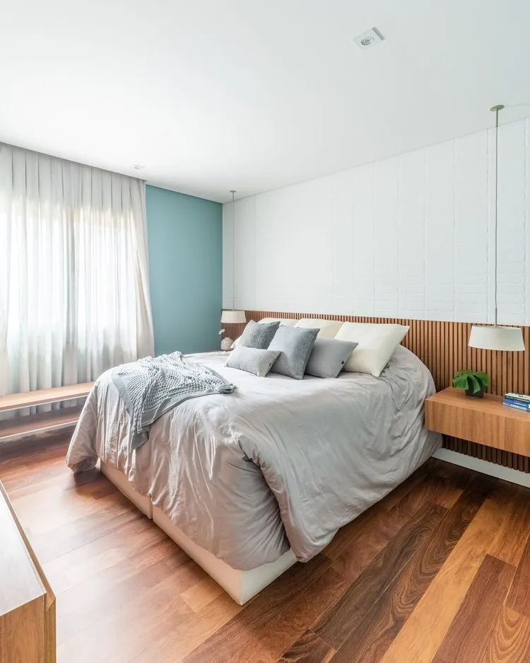 Quarto do casal com cama king size bem confortável. Parede de tijolinhos e mesa de canto suspensa de madeira, com luminárias penduradas no teto.