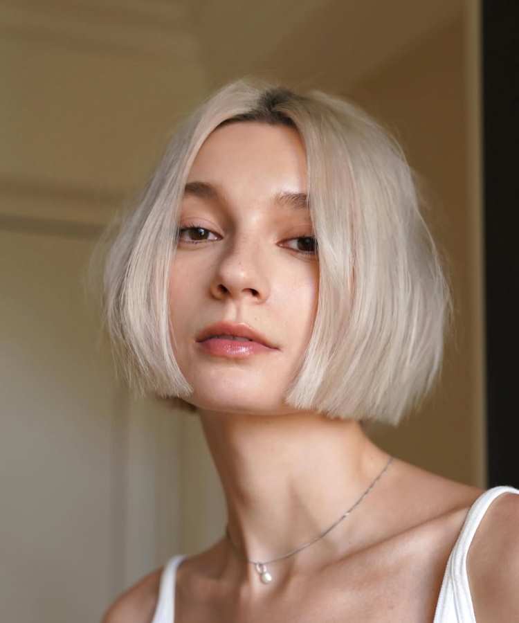 Mulher com cabelo curto estilo bob reto loiro platinado, separação central. Pele clara, maquiagem leve com lábios brilhantes. Ela usa regata branca e colar delicado. Fundo interno suave com iluminação natural, expressando serenidade e elegância no olhar.