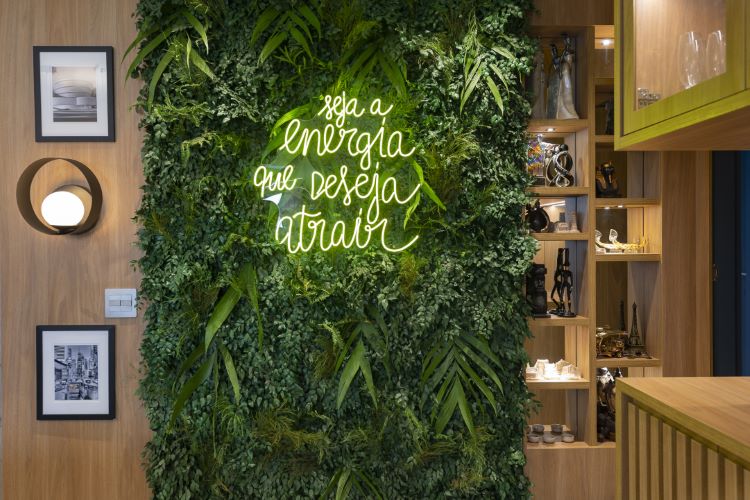 A imagem mostra uma parede verde repleta de plantas, criando um painel natural. No centro, há uma frase em neon verde que diz: "Seja a energia que deseja atrair". Ao lado esquerdo, uma parede de madeira possui duas fotos emolduradas e uma luminária circular moderna. À direita, há uma estante de madeira com nichos decorados com objetos diversos, como esculturas e lembranças de viagem. O ambiente é acolhedor, combinando elementos naturais e modernos, com uma atmosfera leve e inspiradora.
