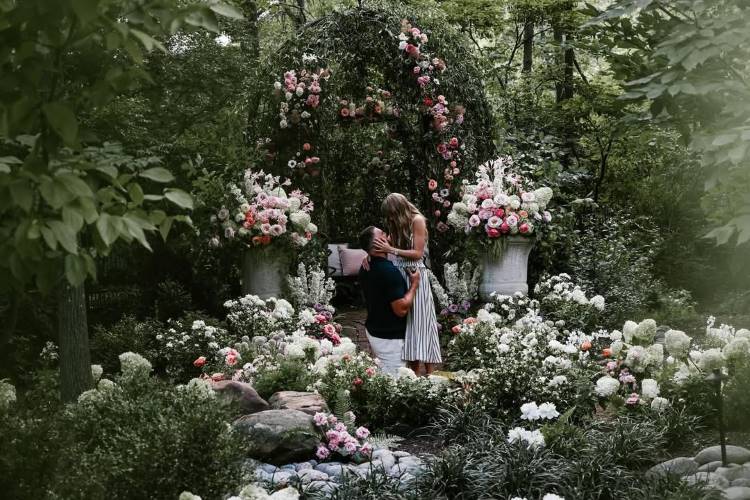 Travis Kelce ajoelhado em um jardim exuberante, com flores e folhagens, pede Taylor Swift em casamento. Eles se beijam sob um arco floral. A cena é romântica e verde, capturando um momento de proposta.