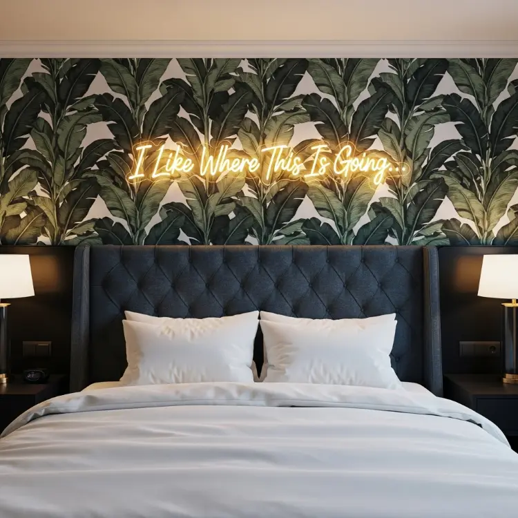 Uma cama com cabeceira estofada azul-marinho. Acima dela, um letreiro de neon dourado com a frase "I Like Where This Is Going.." em uma parede com papel de parede de folhas tropicais verdes e brancas. Duas lâmpadas de mesa estão ao lado da cama.