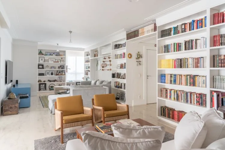Decoração de ambiente integrado de sala de estar com televisão e cantinho da leitura com uma estante repleta de livros, com poltronas marrom e sofá off white, que contribui com a saúde mental