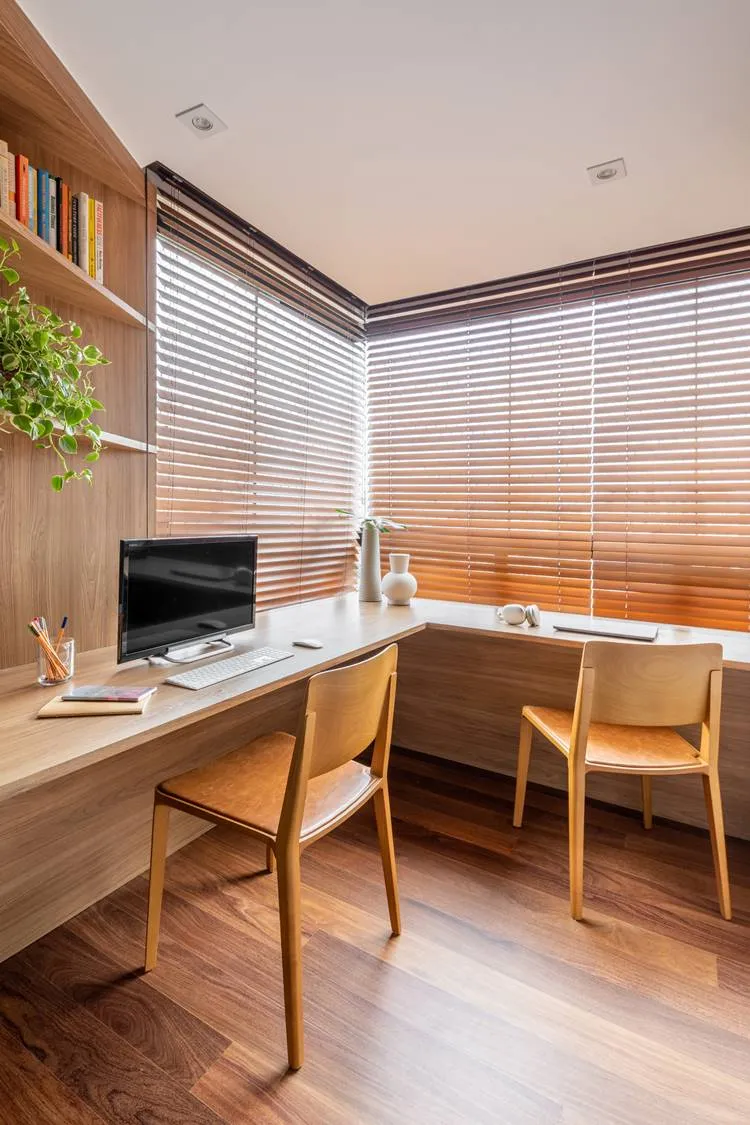 Escritório ou home-office do apê. Mesa em L no canto com persianas. Duas cadeiras de madeira, computador, cadernos, notebook, fone de ouvido e outros objetos.