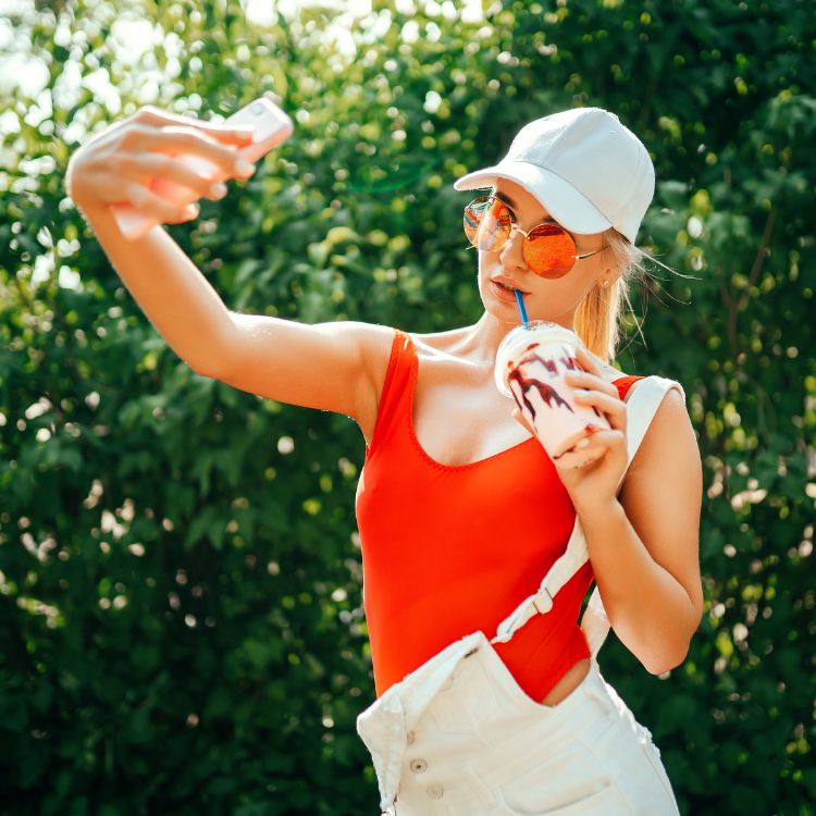 Jovem influencer em ambiente externo, usando óculos de sol e boné branco, tira uma selfie enquanto segura um milkshake. Essa imagem exemplifica o que fazem os influencers: criar conteúdo visual atrativo para engajar seguidores e promover estilo de vida.
