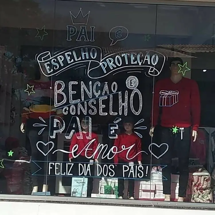 A imagem mostra uma vitrine de loja decorada para o Dia dos Pais. Escrito em branco no vidro, há frases como "Pai é Espelho Proteção Benção e Conselho" e "Pai é Amor Feliz Dia dos Pais!". Dois manequins adultos e dois infantis vestem roupas vermelhas.