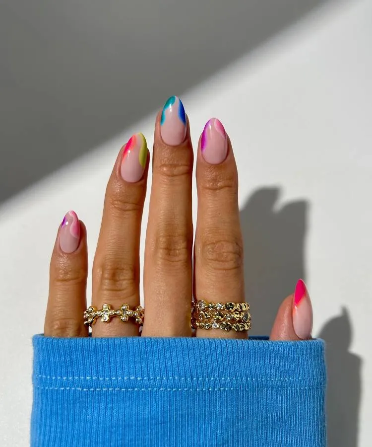A imagem mostra um close-up de uma mão descansando sobre uma superfície, com os dedos estendidos para cima. As unhas estão decoradas com um design único e colorido; cada unha tem uma combinação diferente de cores, criando formas pontiagudas nas pontas, que lembram chamas ou pétalas. As cores incluem azul, rosa, amarelo e verde sobre uma base branca. A pele é de tom claro, e não há indicação da identidade da pessoa. A mão está adornada com vários anéis em diferentes dedos; esses anéis parecem ser de ouro e têm designs intrincados, possivelmente com pedras embutidas.
