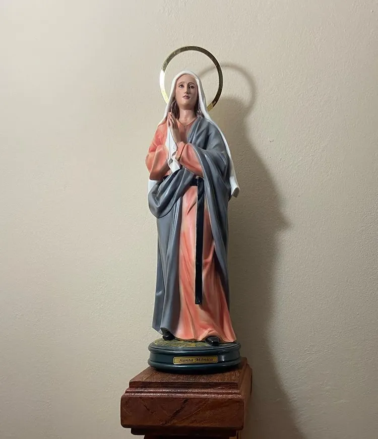 A imagem mostra uma estátua de Santa Mônica. Ela está de pé, com as mãos juntas em oração. A estátua veste um manto cinza sobre uma túnica rosa e um véu branco. Um halo dourado circunda sua cabeça, e a base da estátua possui uma placa com o nome "Santa Mônica". A estátua está colocada sobre um pedestal de madeira.


