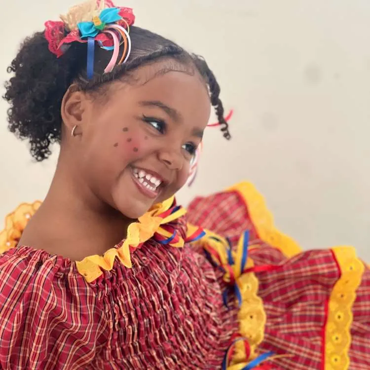 Menina com vestido vermelho e amarelo, usando mini chapéu de palha no cabelo.
