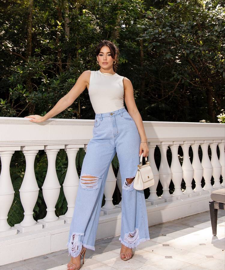 A imagem apresenta uma mulher vestindo uma calça jeans wide leg de lavagem clara com rasgos nos joelhos e na barra, tendência 2025. Ela combina a peça com um top branco ajustado, sandálias de salto e uma bolsa pequena elegante.