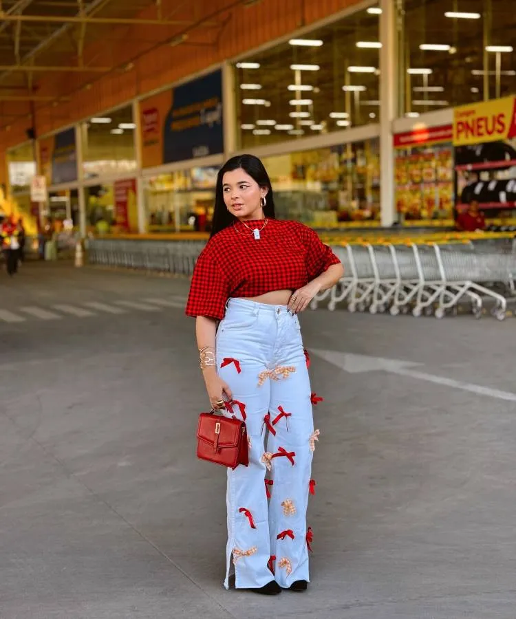 A imagem mostra uma jovem em frente a uma loja. Ela veste uma camisa xadrez vermelha e preta, calça jeans com apliques de laços, e carrega uma bolsa vermelha. O fundo exibe a entrada de uma loja, com carrinhos de compras alinhados.
