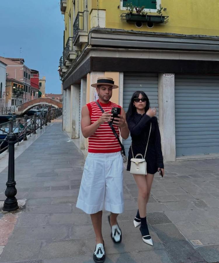 Um homem e uma mulher andam em uma rua estreita de Veneza. O homem tem um chapéu de palha, uma regata listrada de vermelho e branco, e shorts brancos. A mulher usa um minivestido preto, sapatos de salto alto e carrega uma bolsa branca.







