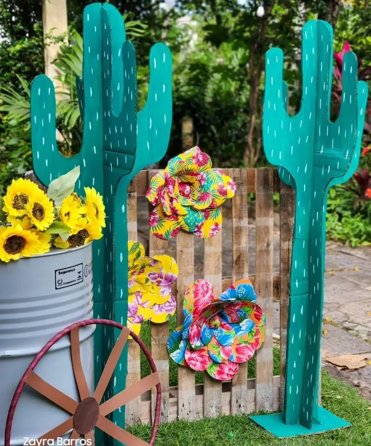 Decoração de festa Junina ao ar livre com dois grandes cactos verdes de madeira. Entre eles, flores de papel coloridas e um palete de madeira. À esquerda, um balde com girassóis e uma roda de carroça.