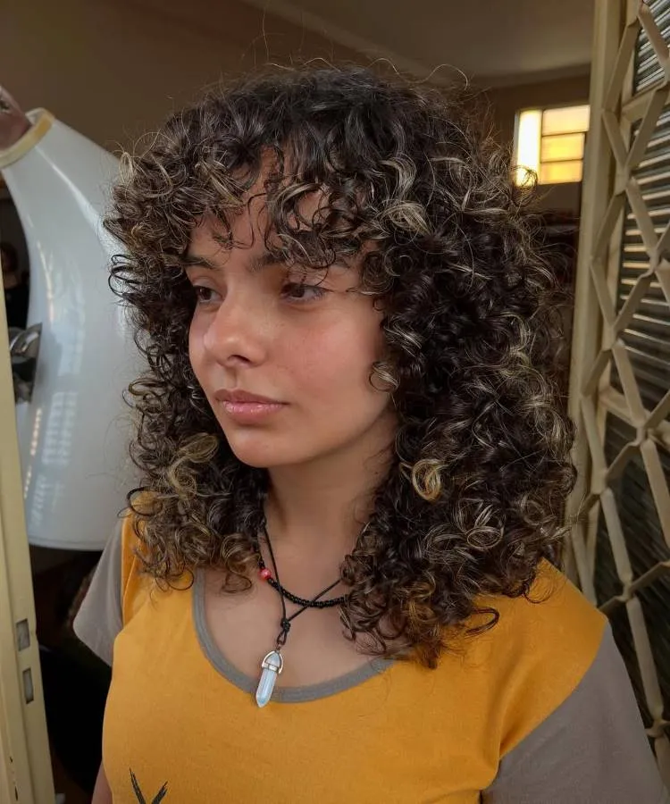 Uma jovem com um corte de cabelo cacheado de comprimento médio, com luzes loiras em meio aos cachos escuros. Ela usa uma blusa amarela e um colar, olhando para o lado com uma expressão pensativa.
