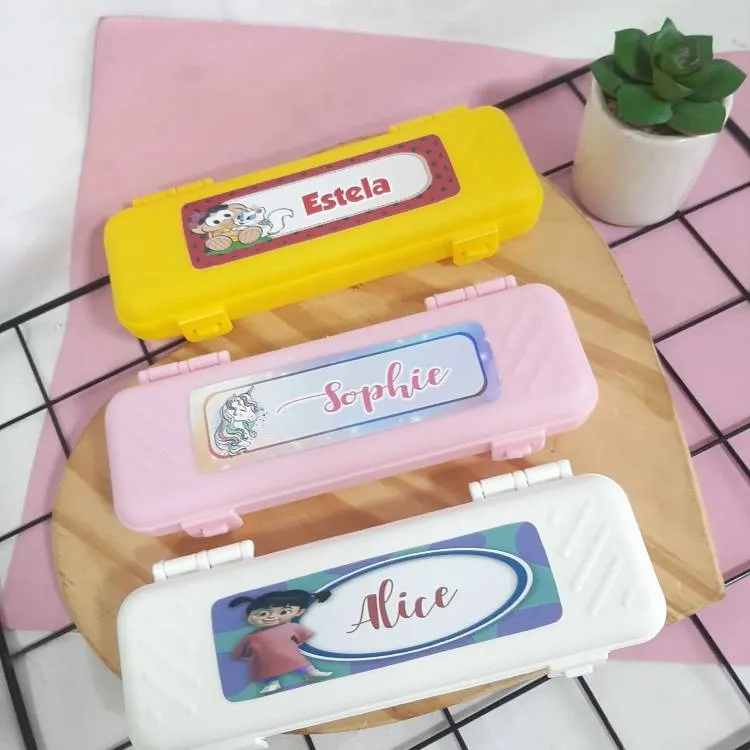 Três estojos de plástico personalizados: um amarelo com a personagem Mônica e "Estela", outro rosa claro com um unicórnio e "Sophie", e um branco com a personagem Boo de Monstros S.A. e "Alice".

