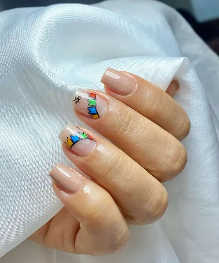Mão com unhas quadradas em tom nude clássico. Duas unhas possuem um desenho de bandeirinhas coloridas: uma com um símbolo de cerquilha e outra com um coração.