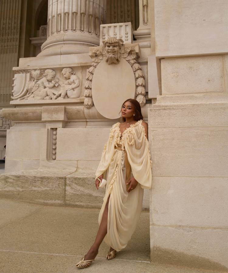 Uma mulher posa elegantemente em frente a um edifício de pedra. Ela usa um vestido creme esvoaçante com mangas compridas e um cinto dourado, sapatos dourados e brincos grandes, olhando para longe da câmera.







