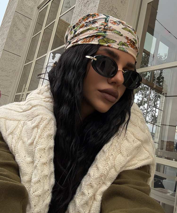 Mulher com cabelo longo e ondulado, usando lenço estampado na cabeça, óculos escuros e casaco bege trançado, posando ao ar livre.