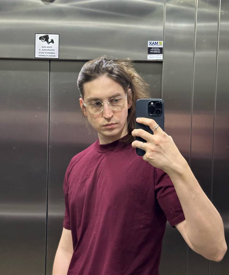 Homem jovem de cabelos castanhos compridos, óculos de grau e camiseta bordô, tirando uma selfie com um celular preto. Ele está dentro de um elevador com portas de metal reflexivas.