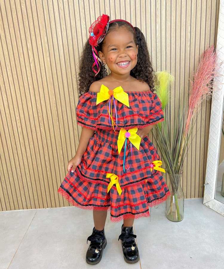 Garotinha usando vestido xadrez vermelho e preto de quadrila, com laços amarelos + adereço no cabelo