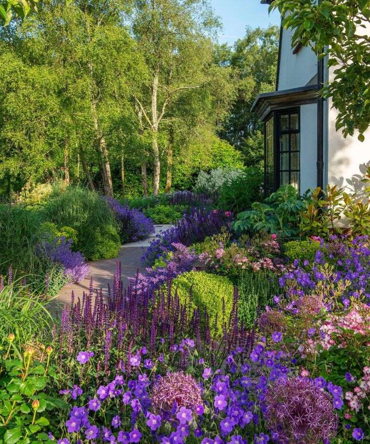 A imagem retrata um exuberante jardim florido, com uma variedade de plantas em tons vibrantes de roxo, lilás e verde. Um caminho sinuoso atravessa o cenário, rodeado por árvores altas, arbustos densos e flores coloridas, criando harmonia natural.