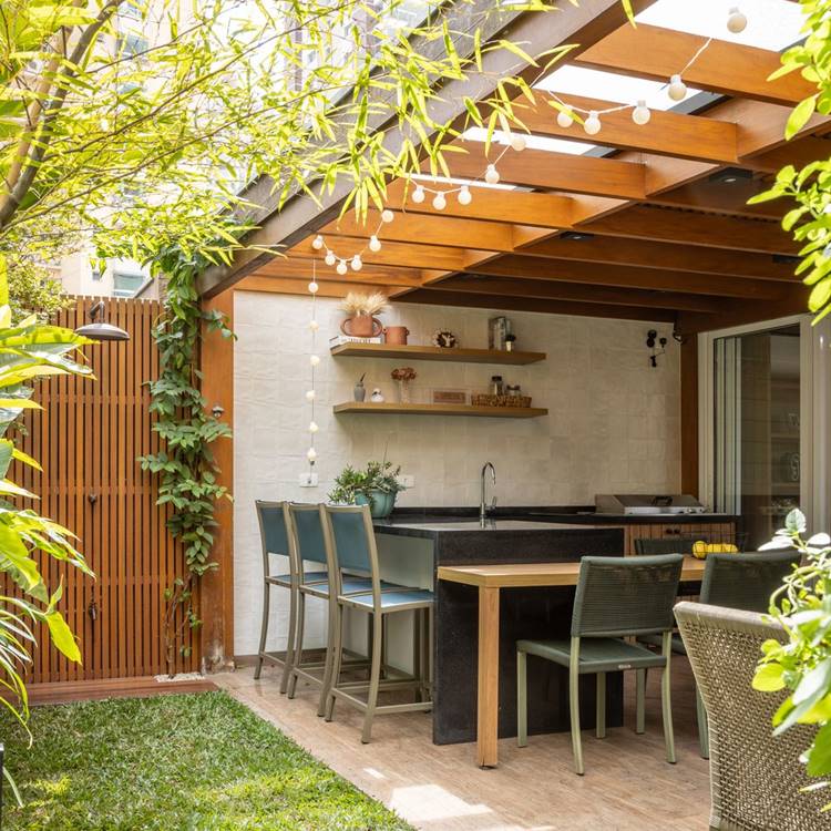 espaço ao ar livre com uma cozinha externa, cadeiras e uma mesa, cercado por vegetação e iluminado por luzes penduradas. O cenário é um jardim ao ar livre com uma cozinha integrada. Há uma estrutura de madeira acima do espaço, com várias lâmpadas penduradas que provavelmente proporcionam uma iluminação suave à noite. A cozinha tem um balcão de pedra preta, armários embutidos e prateleiras de madeira na parede que exibem vários itens de cozinha. Existem quatro cadeiras em tons de verde azulado e cinza em volta da mesa que combina com o balcão da cozinha. O chão é parcialmente coberto por grama verde e piso de pedra ou concreto. Uma cerca de madeira marrom e vegetação circunda o espaço