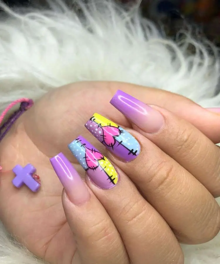 Mão com unhas longas no formato bailarina. As unhas laterais têm um degradê roxo. As unhas do meio são decoradas com um padrão de retalhos coloridos em tons de roxo, azul e amarelo, com corações.