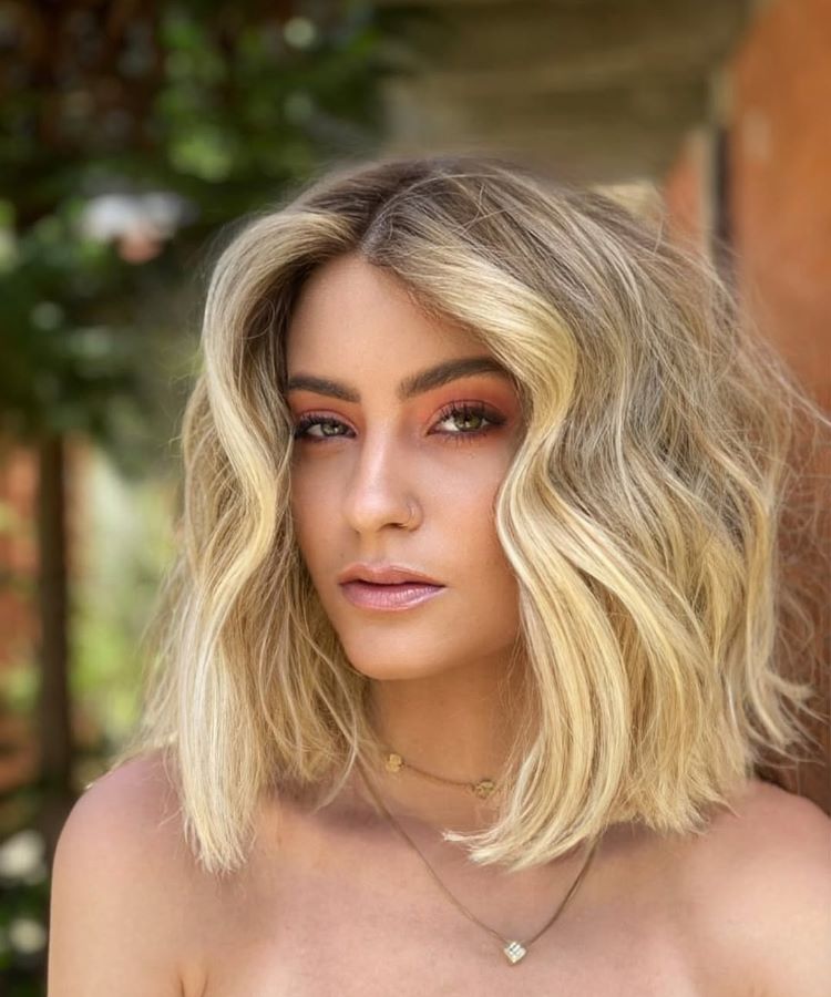Mulher com cabelos curtos e ondulados, exibindo uma das tendências modernas de cores de cabelo: loiro platinado com nuances douradas. Sua maquiagem destaca olhos com tons de coral e pele iluminada. Fundo externo com vegetação desfocada.