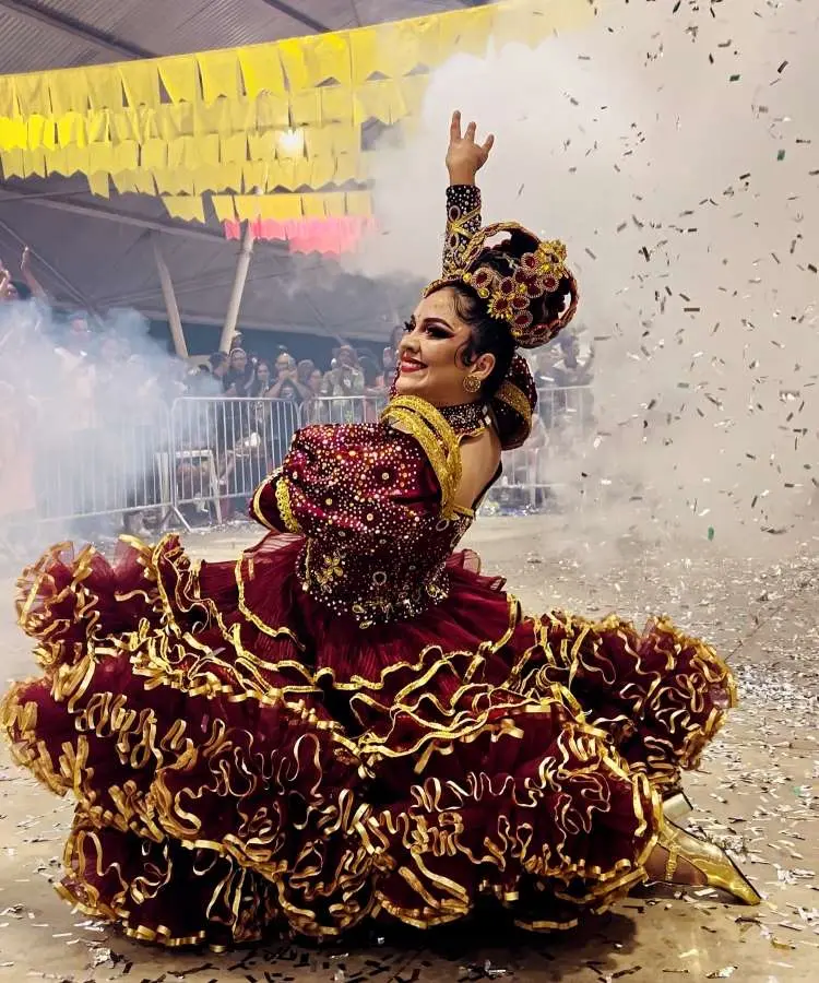 Imagem de uma rainha da quadrilha em festa junina, vestindo um traje vermelho e dourado exuberante, com saia rodada e detalhes ricos. Ela posa alegremente com um braço levantado, em meio a fumaça e confetes, celebrando a cultura popular brasileira.