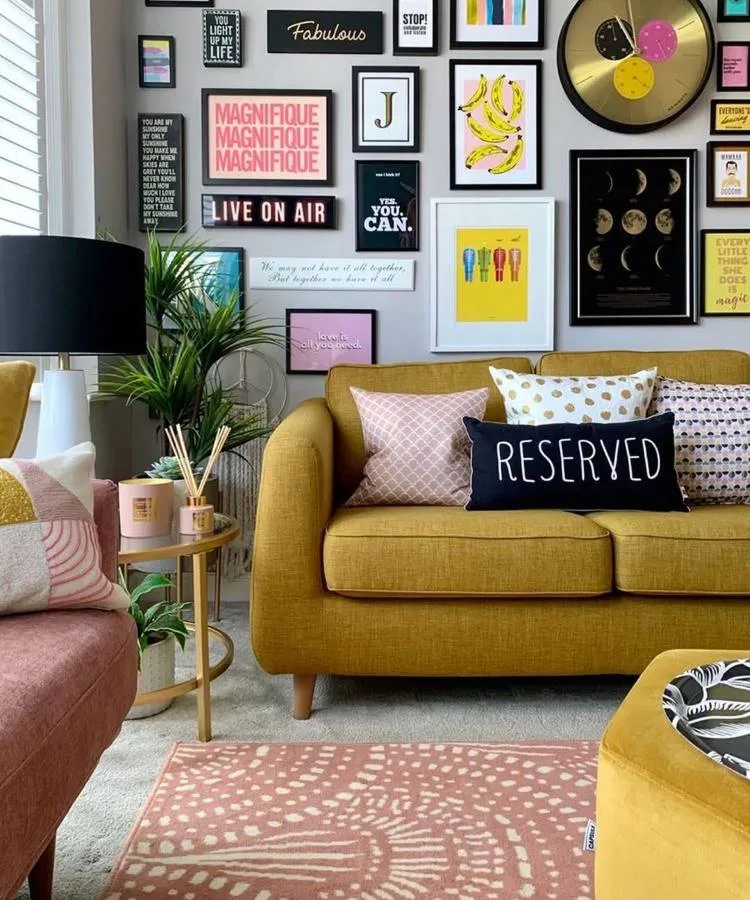 Uma sala vibrante com sofá mostarda e almofadas para sala decorativas, incluindo uma com a palavra "RESERVED". A parede cinza é coberta por uma galeria de quadros coloridos e artísticos. Um tapete rosa com estampa geométrica e uma mesa lateral dourada completam o estilo eclético e divertido.







