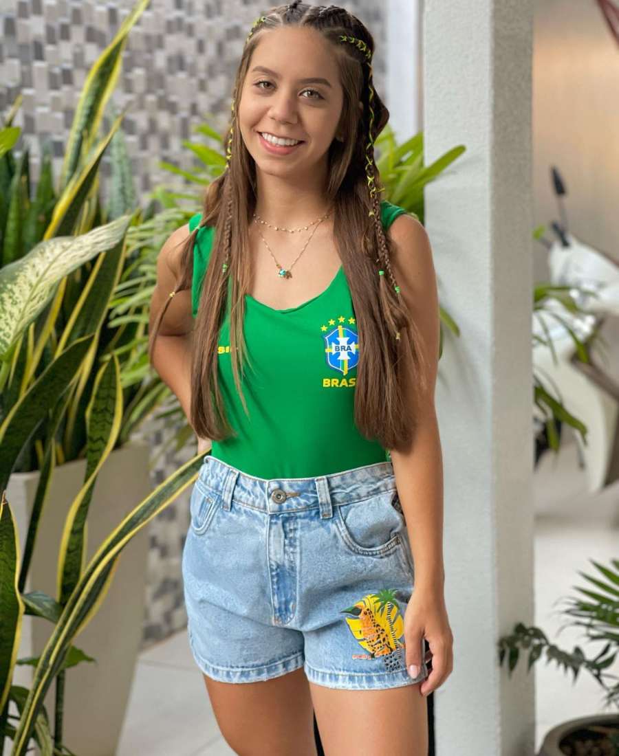 Imagem com fundo de parede e plantas. No centro, mulher usando roupa copa do mundo: body verde do Brasil + short jeans.