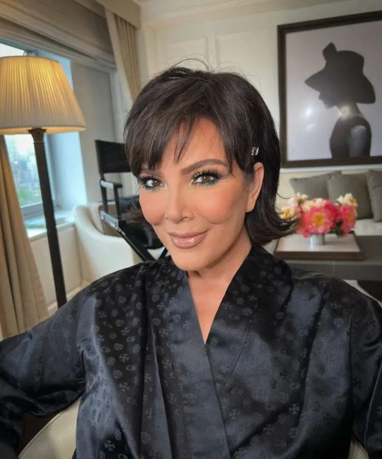 Kris Jenner, de robe preto, sorri levemente para a câmera. Seu cabelo escuro e curto está penteado para o lado, e sua maquiagem realça os olhos com um toque de verde.