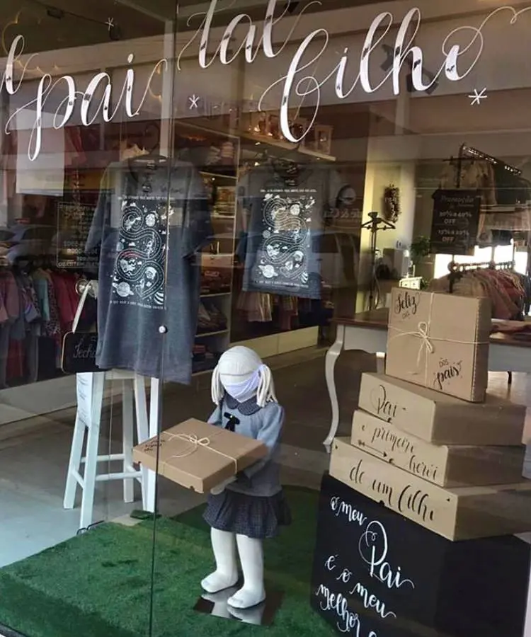 A imagem mostra uma vitrine de loja com a frase "tal pai tal filho" escrita em caligrafia no vidro. Há camisetas expostas, uma boneca manequim infantil segurando uma caixa de presente e uma pilha de caixas com mensagens sobre o Dia dos Pais.